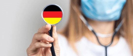 Doctor german care lucreaza in sistemul sanitar german femeie care se află în plan secund cu o mască sanitară pe față și cu stetoscopul în ambele urechi, iar în plan principal este continuarea stetoscopului pe care se afla colorat steagul Germaniei cu negru, roșu și galben.