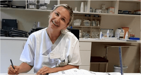 Asistentă Medicală îmbrăcată în alb, care se află la un birou într-un cabinet medical din Germania