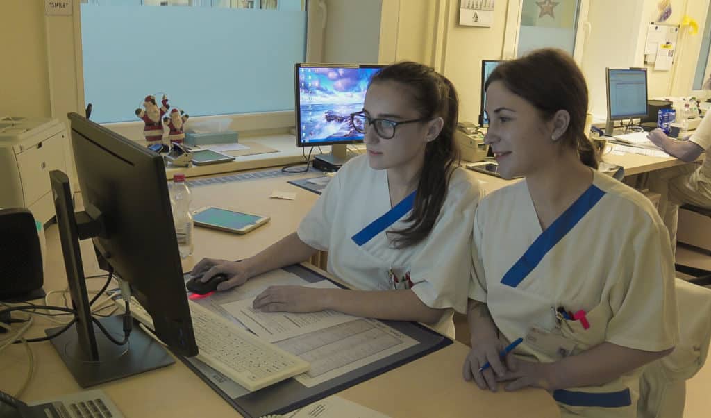 Cabinet medical în Germania în care se află 2 asistente îmbrăcate în alb care lucrează la un calculator