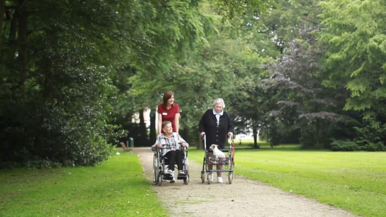 Curte a unei reședințe de seniori MediClub Germania în care se află o asistentă medicală care îîmpinge un cărucior cu rotile în care se află o bătrânp și lângă pe alee tot mergând se află o bătrână care împinge un cărucior cu un cățel